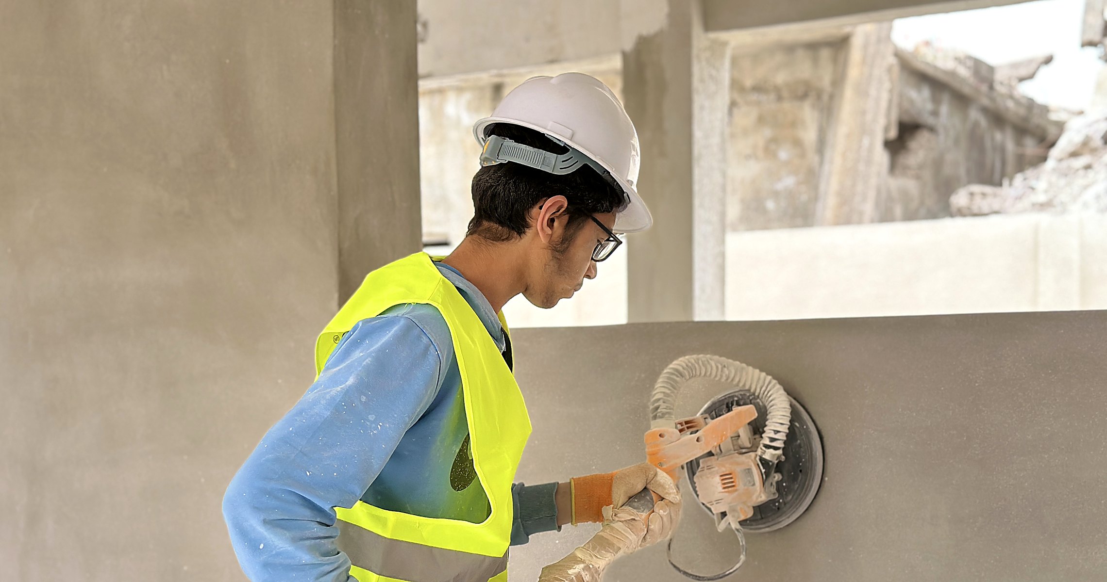 An Uncle T builder in a hard hat resurfacing a classroom wall, war-damaged structure still visible through the opening beyond