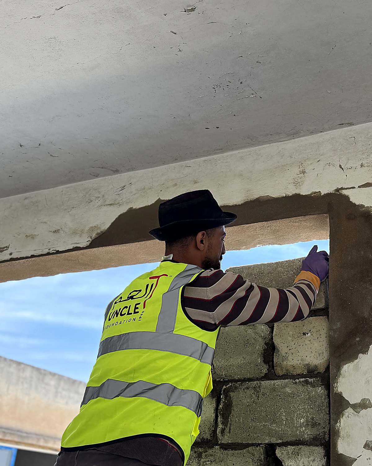 An Uncle T builder in a branded high-visibility vest rebuilding a doorway, open sky beyond the wall